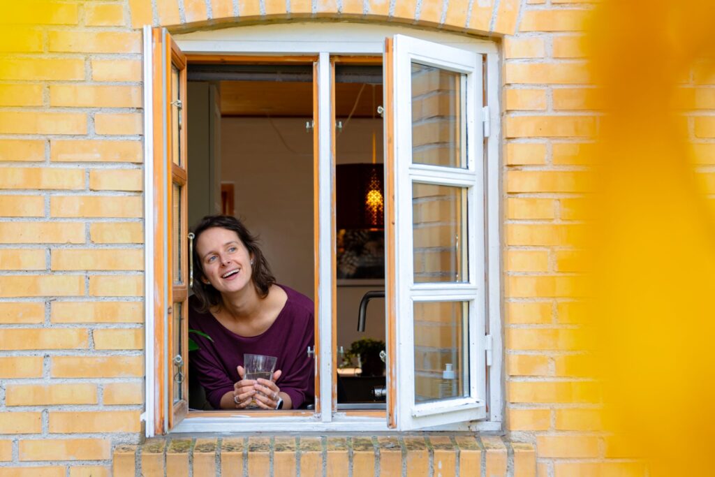 Marlies is looking out of a window with a cup of tea in her hand. She is smiling.